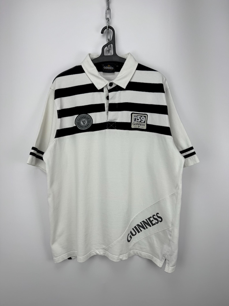 May include: White polo shirt with black stripes across the chest and sleeves. The shirt displays the Guinness logo and the word "Guinness" printed diagonally. The shirt is suspended from a black hanger. The shirt is a classic design.