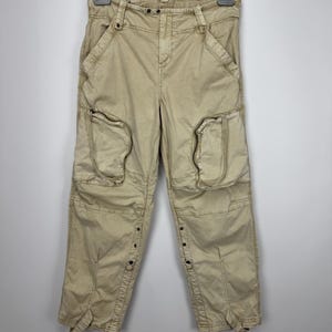 May include: Tan cargo pants with multiple pockets and button details. The pants are hanging on a metal hanger. The pants have a relaxed fit and are made of a durable fabric. The design includes large pockets on the thighs and knees.