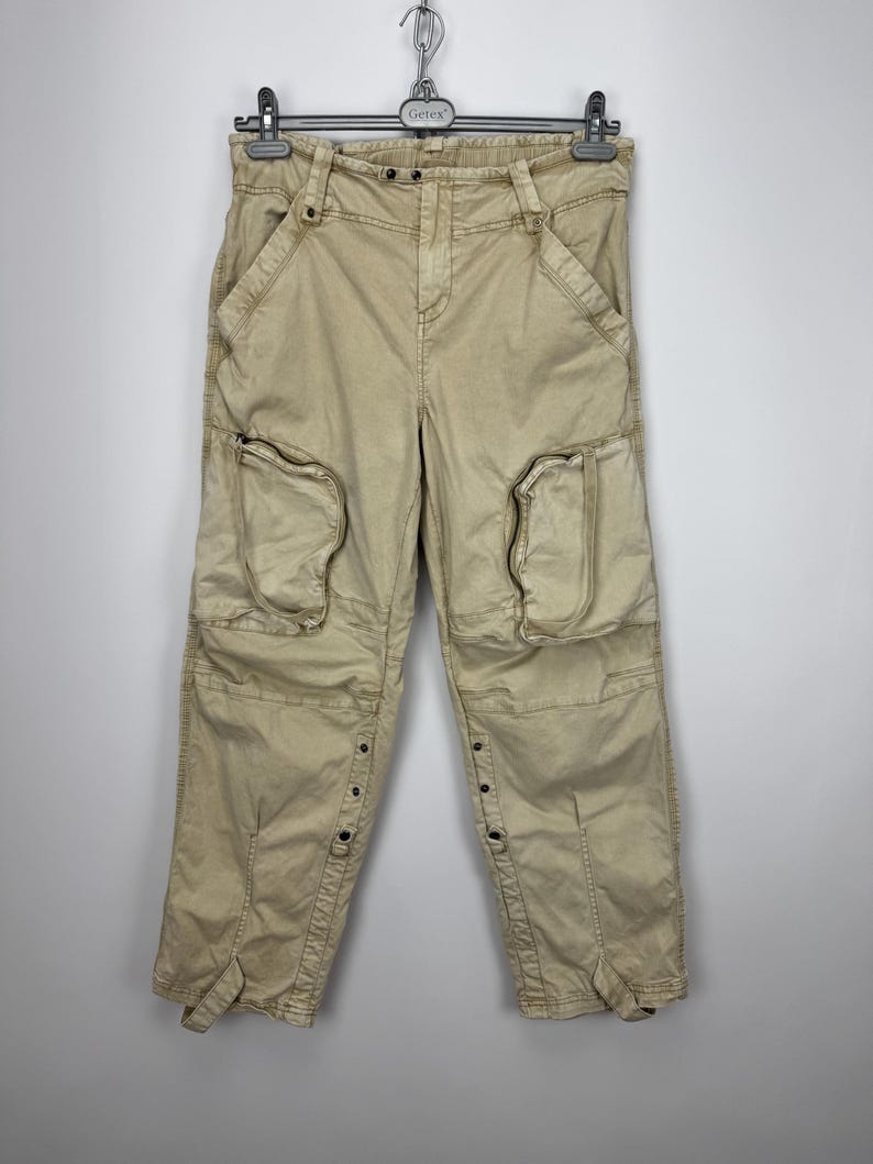 May include: Tan cargo pants with multiple pockets and button details. The pants are hanging on a metal hanger. The pants have a relaxed fit and are made of a durable fabric. The design includes large pockets on the thighs and knees.