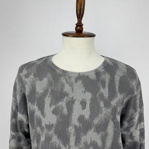 May include: A grey crew neck sweater with a tie-dye pattern. The sweater is displayed on a mannequin. The fabric has a textured appearance, and the pattern features shades of grey and white.