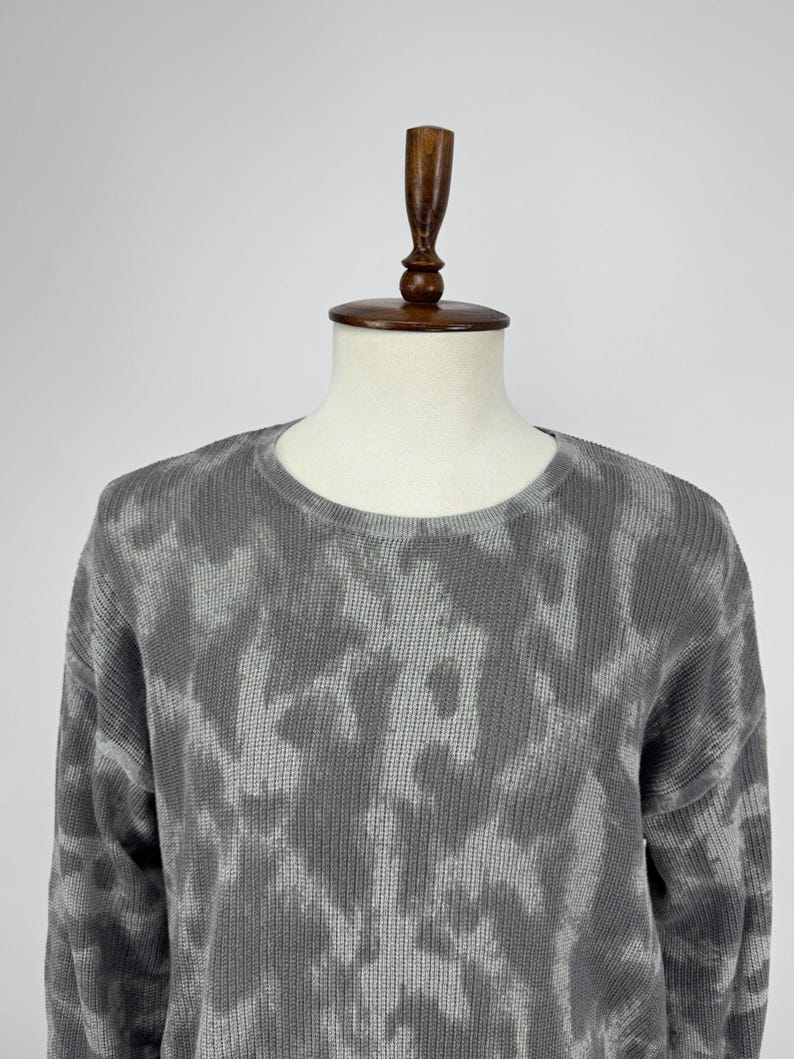 May include: A grey crew neck sweater with a tie-dye pattern. The sweater is displayed on a mannequin. The fabric has a textured appearance, and the pattern features shades of grey and white.