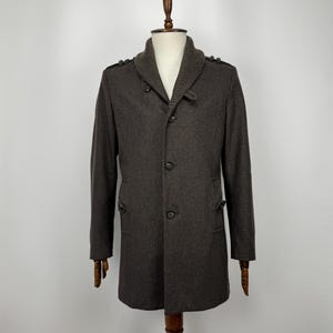 May include: A dark brown coat featuring a ribbed shawl collar, a three-button front closure, and shoulder epaulets. The coat includes two side pockets with buttoned flaps and long sleeves, displayed on a mannequin against a plain background.