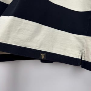 Puede incluir: Primer plano de una camiseta a rayas azul marino y blanca. La camiseta presenta gruesas rayas horizontales y una peque&ntilde;a etiqueta dorada con un emblema de arpa. La tela parece ser un material de punto suave.