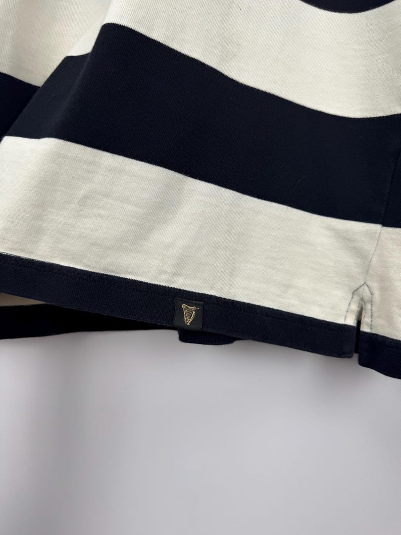Puede incluir: Primer plano de una camiseta a rayas azul marino y blanca. La camiseta presenta gruesas rayas horizontales y una peque&ntilde;a etiqueta dorada con un emblema de arpa. La tela parece ser un material de punto suave.