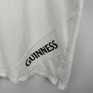 May include: White t-shirt featuring the black word "GUINNESS" printed on the lower right. The shirt has a diagonal seam detail. The material looks soft and breathable.