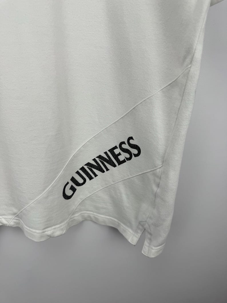 May include: White t-shirt featuring the black word "GUINNESS" printed on the lower right. The shirt has a diagonal seam detail. The material looks soft and breathable.