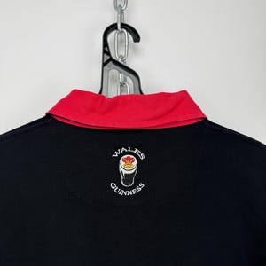 Może przedstawiać: Czarna koszulka polo z czerwonym kołnierzem. Koszulka ma wyszywane logo "Wales Guinness" z tyłu. Koszulka wisi na czarnym wieszaku i srebrnym łańcuchu.