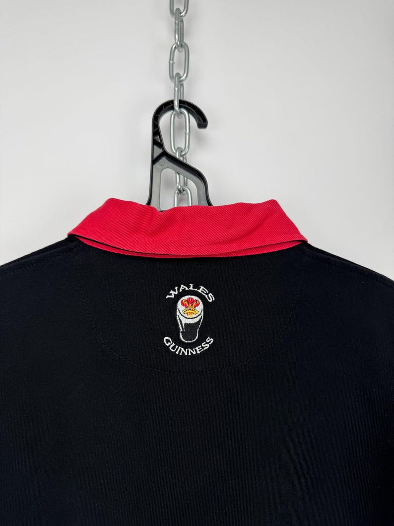 Może przedstawiać: Czarna koszulka polo z czerwonym kołnierzem. Koszulka ma wyszywane logo "Wales Guinness" z tyłu. Koszulka wisi na czarnym wieszaku i srebrnym łańcuchu.