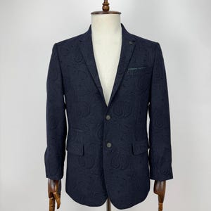 Op de afbeelding: Marineblauwe blazer met een paisley patroon. De blazer heeft een reverskraag, twee voorzakken en een borstzak. Het wordt getoond op een houten mannequin. De blazer heeft twee knopen en een kleine decoratieve knoop op de revers.