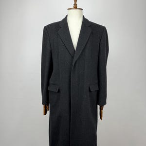 Op de afbeelding: Een donkergrijze, lange wollen jas, getoond op een houten mannequin. De jas heeft een revers, twee voorzakken en een klassiek, getailleerd silhouet. De jas is knielang en heeft een ontwerp met één rij knopen.