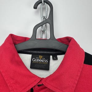 Może przedstawiać: Koszulka do rugby w czerwono-czarne paski z czerwonym kołnierzem i logo Guinness. Koszulka wisi na czarnym wieszaku i srebrnym łańcuchu. Etykieta głosi "Official Merchandise".