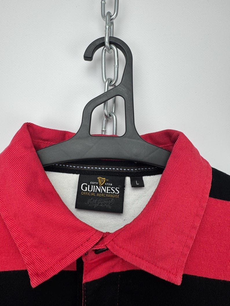 Może przedstawiać: Koszulka do rugby w czerwono-czarne paski z czerwonym kołnierzem i logo Guinness. Koszulka wisi na czarnym wieszaku i srebrnym łańcuchu. Etykieta głosi "Official Merchandise".