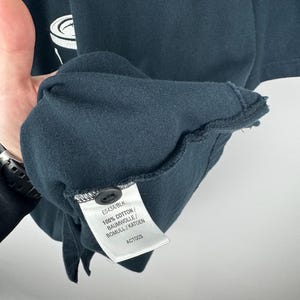 Może przedstawiać: Zbliżenie na ciemnoniebieską koszulkę z małym czarnym guzikiem i metką. Na metce widnieje napis "E543A/BLK" oraz "100% COTTON / BAUMWOLLE / BOMULL / KATOEN". Koszulka ma biało-zielony wz&oacute;r.