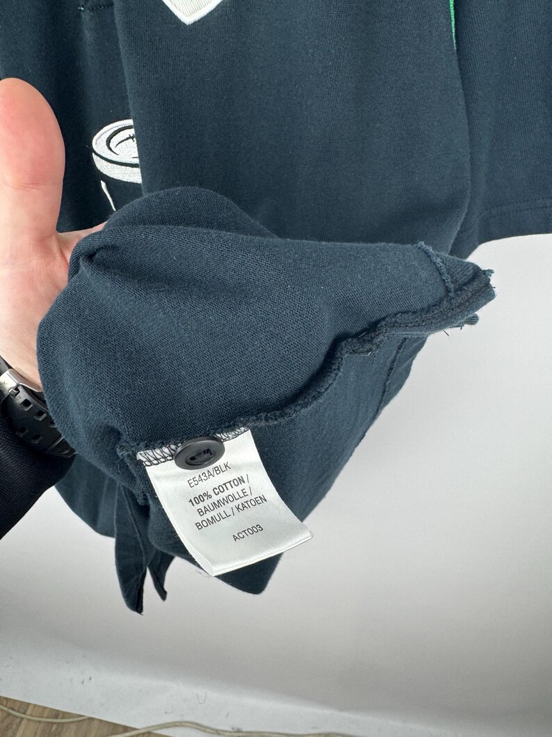 Może przedstawiać: Zbliżenie na ciemnoniebieską koszulkę z małym czarnym guzikiem i metką. Na metce widnieje napis "E543A/BLK" oraz "100% COTTON / BAUMWOLLE / BOMULL / KATOEN". Koszulka ma biało-zielony wz&oacute;r.