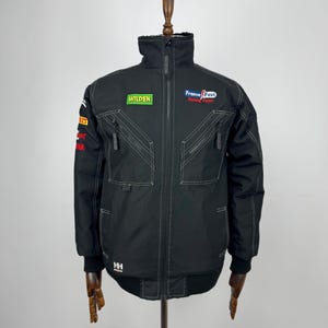 Puede incluir: Chaqueta negra con costuras blancas y varios parches, incluyendo "WILDEN" y "Frame Fast Racing Team". La chaqueta tiene un cuello alto, una cremallera completa y múltiples bolsillos. La chaqueta se exhibe en un maniquí de madera.