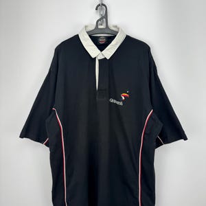 Puede incluir: Polo negro de manga corta con cuello blanco y ribete rojo y blanco. El logotipo de Guinness est&aacute; bordado en el pecho. La camisa est&aacute; colgada de una percha de metal.