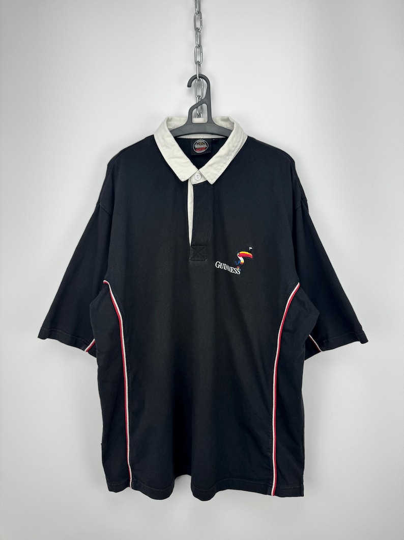 Puede incluir: Polo negro de manga corta con cuello blanco y ribete rojo y blanco. El logotipo de Guinness est&aacute; bordado en el pecho. La camisa est&aacute; colgada de una percha de metal.