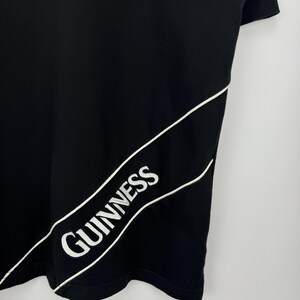 Puede incluir: Camiseta negra con la palabra "Guinness" en blanco, contorneada en blanco, en diagonal en la parte delantera. La camiseta tiene cuello redondo y mangas cortas. El logo de Guinness es un dise&ntilde;o cl&aacute;sico.