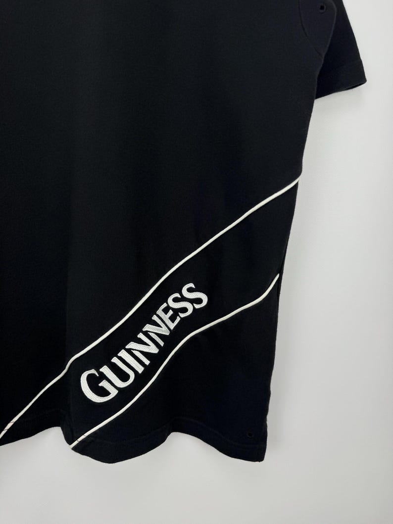 Puede incluir: Camiseta negra con la palabra "Guinness" en blanco, contorneada en blanco, en diagonal en la parte delantera. La camiseta tiene cuello redondo y mangas cortas. El logo de Guinness es un dise&ntilde;o cl&aacute;sico.