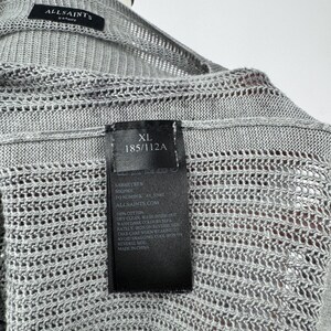 May include: A light grey XL AllSaints knit cardigan. The open-weave design is visible. A black tag displays the size, care instructions, and the brand's website. The cardigan is made of 100% cotton. The size is 185/112A.