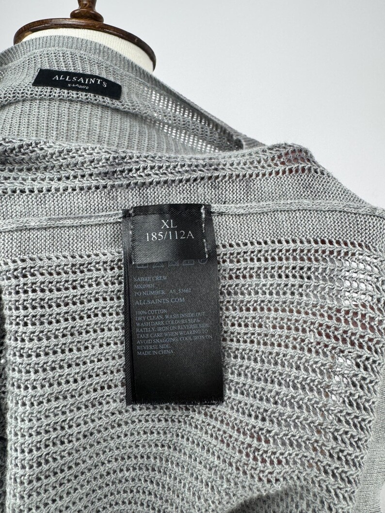 May include: A light grey XL AllSaints knit cardigan. The open-weave design is visible. A black tag displays the size, care instructions, and the brand's website. The cardigan is made of 100% cotton. The size is 185/112A.