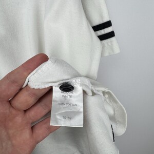 May include: White long-sleeved shirt with a black stripe across the chest and black stripes on the sleeve cuffs. The shirt has a tag that reads "AL90092 WHITE 100% COTTON 70169". The shirt has a black button.