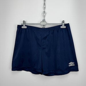 May include: Navy blue athletic shorts featuring a subtle tonal diamond pattern throughout the fabric. The shorts have an elastic waistband and a white embroidered Umbro logo on the lower right leg. The garment is suspended from a metal hanger.