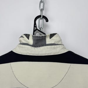 Puede incluir: Una camiseta de rugby a rayas negras y crema, colgada de una percha negra y una cadena plateada. El cuello presenta un dise&ntilde;o de la Union Jack. El dise&ntilde;o de la camiseta incluye rayas horizontales y un cuello blanco.