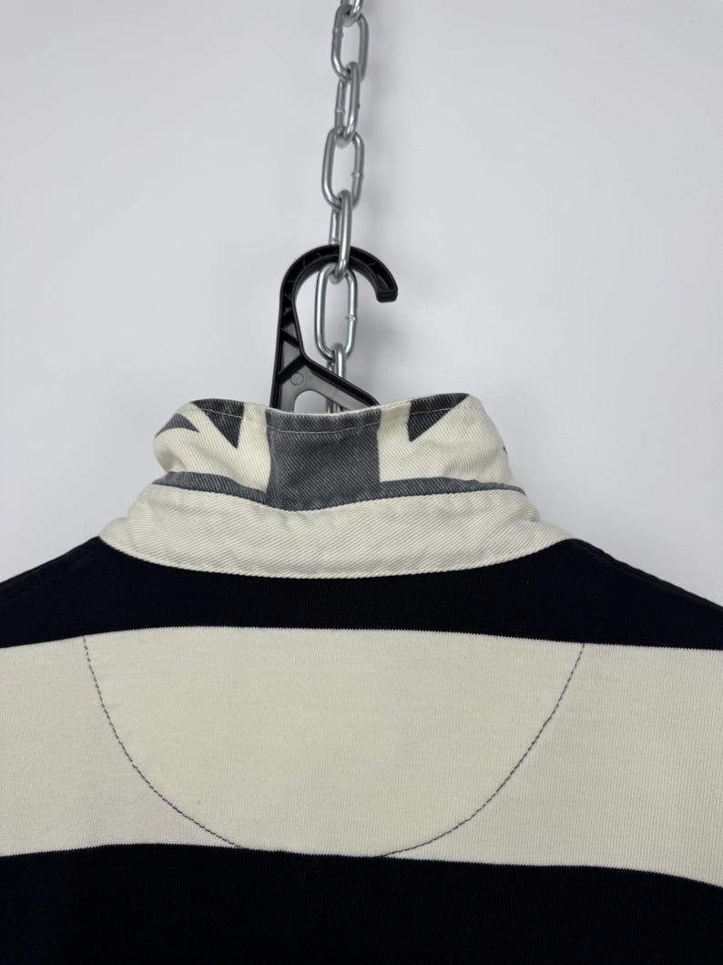 Puede incluir: Una camiseta de rugby a rayas negras y crema, colgada de una percha negra y una cadena plateada. El cuello presenta un dise&ntilde;o de la Union Jack. El dise&ntilde;o de la camiseta incluye rayas horizontales y un cuello blanco.