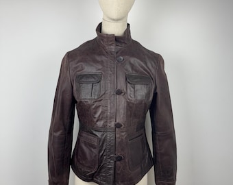 Könnte beinhalten: Dunkelbraune Lederjacke mit Stehkragen und Knopfverschluss. Die Jacke hat zwei Brusttaschen und zwei untere Taschen. Die Jacke wird auf einer Schaufensterpuppe präsentiert.