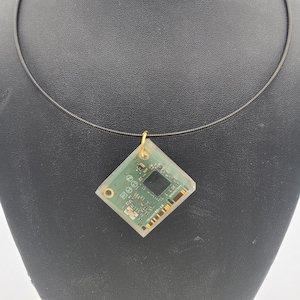 May include: A unique necklace featuring a square pendant made from a green circuit board encased in clear resin. The pendant is suspended from a thin, black cord. The pendant is attached to a gold-colored bail.