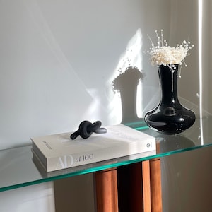 Puede incluir: Una mesa de vidrio con un jarrón negro lleno de flores blancas. Una escultura negra anudada se encuentra sobre un libro blanco titulado "AD at 100".