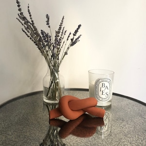 Puede incluir: Una escultura decorativa de nudo de terracota sobre una mesa con espejo, junto a un jarrón de cristal con lavanda y una vela encendida en un tarro de cristal. La vela tiene una etiqueta en blanco y negro. La mesa tiene un marco de metal negro.