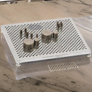 May include: A silver metal tray with a grid pattern and a set of metal cylinders and rods. The tray is on a light brown countertop.