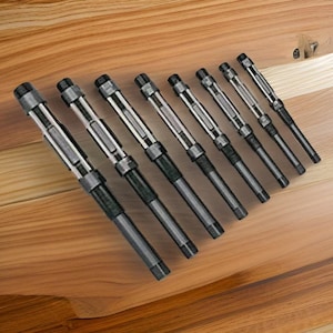 May include: An angled overhead shot displays eight reamers, a type of cutting tool, arranged on a wooden surface. The reamers have a dark gray body with silver-colored sections and black ends. The wooden surface has a light brown color with visible wood grain.