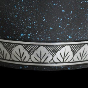 Pode incluir: Close-up de um pote de cer&acirc;mica preta com salpicos azuis. O pote apresenta uma faixa decorativa prateada com um desenho de folhas. A borda do pote &eacute; vis&iacute;vel contra um fundo preto.