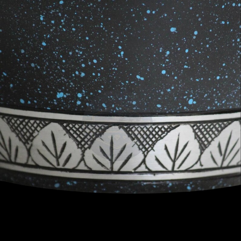 Pode incluir: Close-up de um pote de cer&acirc;mica preta com salpicos azuis. O pote apresenta uma faixa decorativa prateada com um desenho de folhas. A borda do pote &eacute; vis&iacute;vel contra um fundo preto.