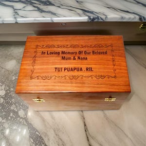 Pode incluir: Uma caixa retangular de madeira com tampa articulada e fechos dourados. A caixa &eacute; gravada com as palavras "In Loving Memory Of Our Beloved Mum & Nana" e "TUI PUAPUA.RIL" em uma fonte decorativa.