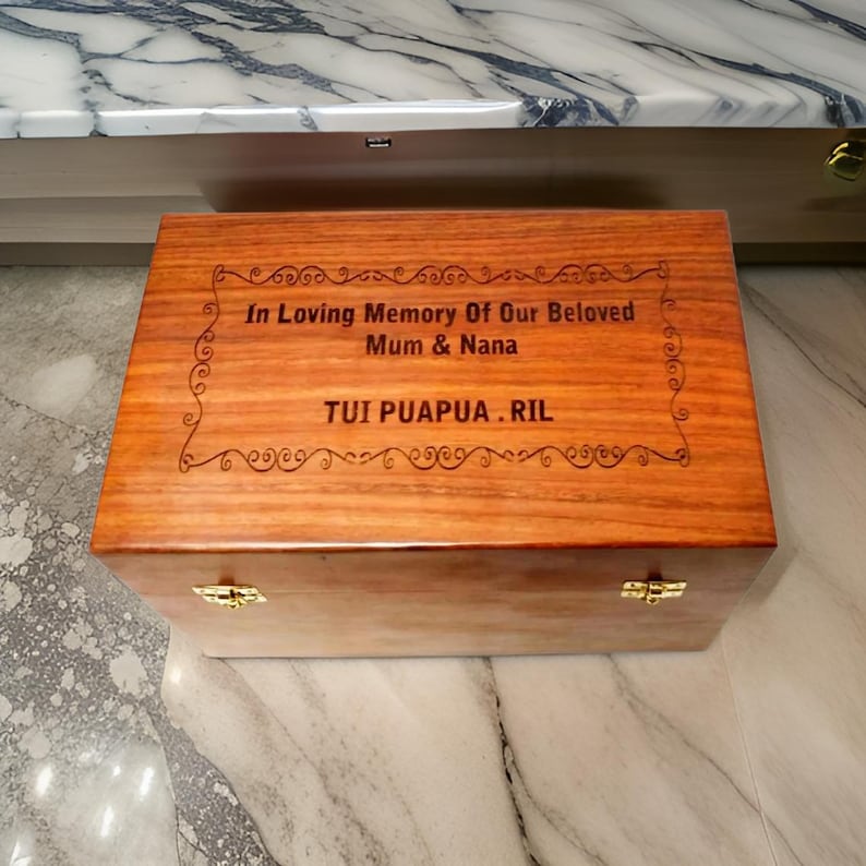 Pode incluir: Uma caixa retangular de madeira com tampa articulada e fechos dourados. A caixa &eacute; gravada com as palavras "In Loving Memory Of Our Beloved Mum & Nana" e "TUI PUAPUA.RIL" em uma fonte decorativa.