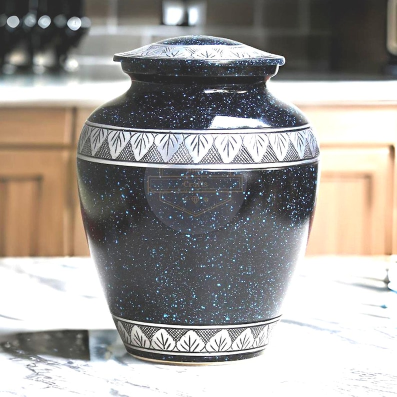 Pode incluir: Uma urna de crema&ccedil;&atilde;o preta salpicada com um design de folhas prateadas na parte superior e inferior. A urna tem uma tampa a condizer e um emblema decorativo na frente. Feita de um material dur&aacute;vel.
