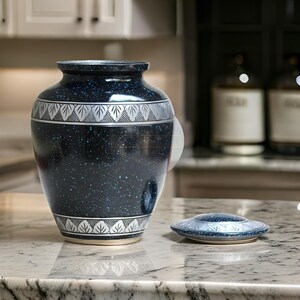 Pode incluir: Uma urna funer&aacute;ria azul escuro com um padr&atilde;o salpicado e um design de folhas prateadas. A urna tem uma tampa a condizer e &eacute; feita de cer&acirc;mica. A urna tem aproximadamente 25 cm de altura.