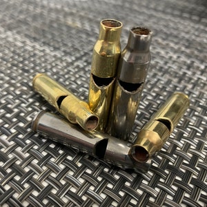 May include: Five brass and silver bullet casings that have been modified into whistles. The whistles are arranged in a pile on a woven black and white surface.