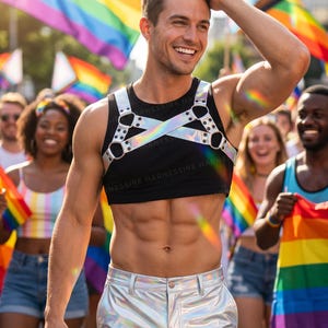 May include: A person wearing a black crop top with a silver harness and silver pants. The person is smiling and surrounded by people holding rainbow flags. The image is taken outdoors on a sunny day.