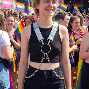 May include: A person wearing a black crop top and denim skirt, with a silver harness featuring iridescent straps and a chain. The background shows a Pride event with rainbow flags and a crowd.