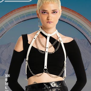 May include: A white leather harness with silver chains and black accents. The harness is worn over a black off-the-shoulder top. The image includes the text "HARNESSIRE" and size options: XS, S, M, L, XL, and 2XL. A rainbow is in the background.