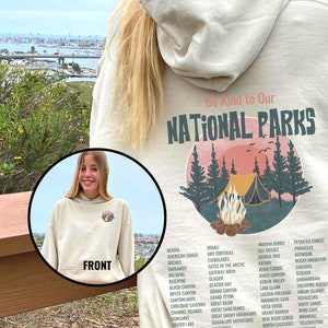 May include: A beige hoodie with a graphic print of a campfire scene in a forest. The text "Be Kind to Our National Parks" is printed above the graphic. A list of national parks is printed below the graphic.