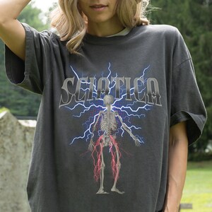 May include: A charcoal gray short-sleeve t-shirt featuring a graphic of a human skeleton with red nerve pathways extending down the legs. Behind the skeleton are blue lightning bolts and the word SCIATICA in a stylized white font.