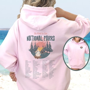 May include: Pink hoodie with a graphic design of a campfire in a forest with the text "Be Kind to Our National Parks" and a list of national parks printed below the graphic.