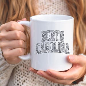 Może przedstawiać: Biały ceramiczny kubek z napisem "NORTH CAROLINA" w szarym kamuflażu. Kubek ma zakrzywiony uchwyt i jest trzymany przez osobę ubraną w kremowy sweter.