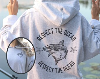 May include: A light gray hoodie with a shark graphic and the text "Respect the Ocean." The design includes a shark illustration, starfish, and the phrase repeated around the shark. A smaller shark logo is on the front.